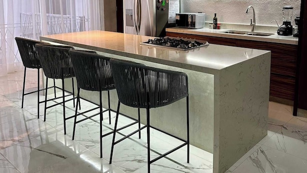 Uma cozinha moderna com ilha central, banquetas, fogão embutido e uma janela grande com cortinas leves.