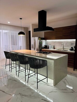 A modern kitchen with a central island, bar stools, a built-in stove, and a large window with sheer curtains.