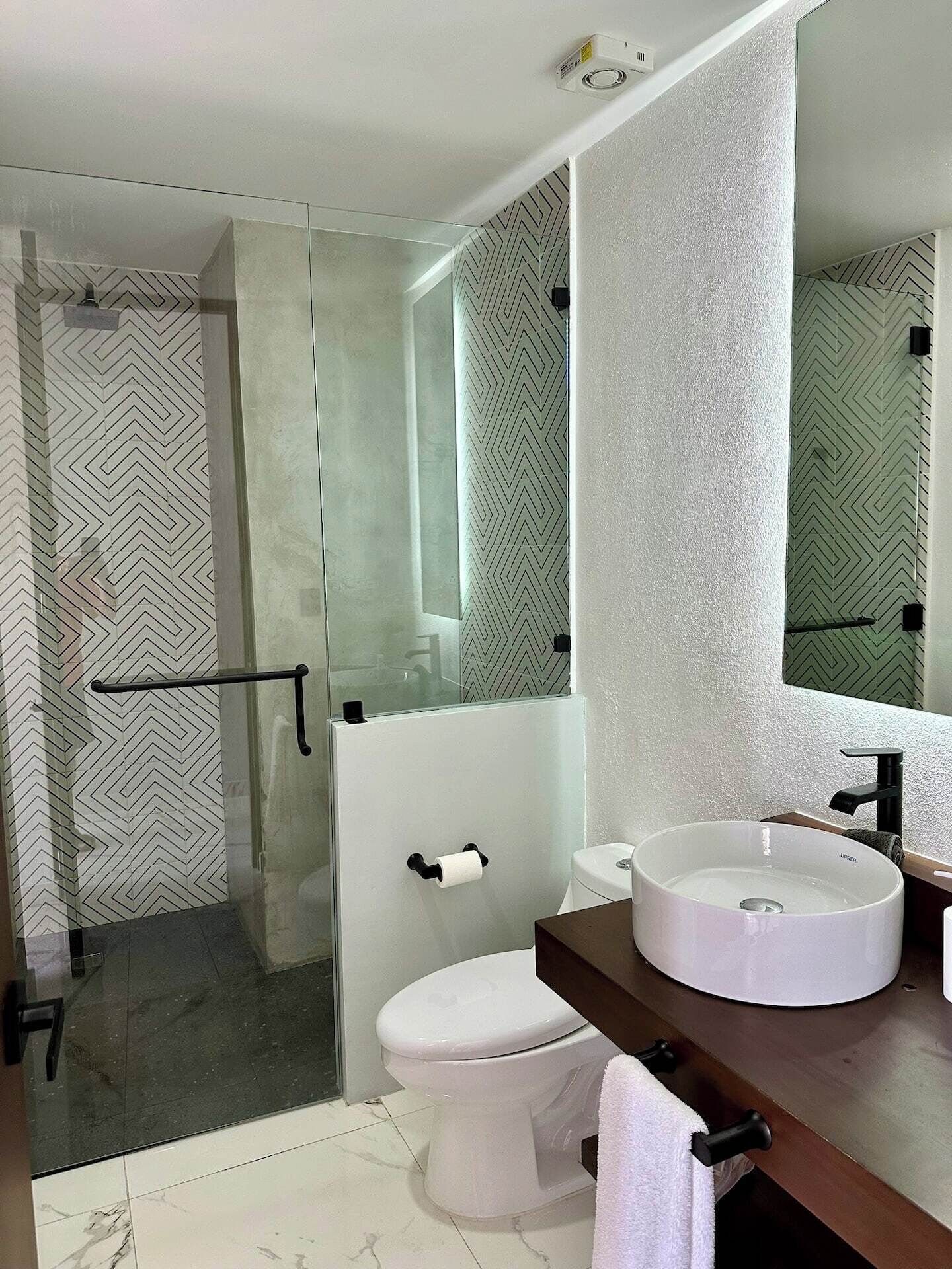 Un baño moderno con ducha envuelta en vidrio, inodoro blanco y lavamanos redondo con espejo superior.