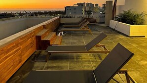 Terraço no telhado com espreguiçadeiras, painel de madeira e vista para o horizonte da cidade ao pôr do sol.
