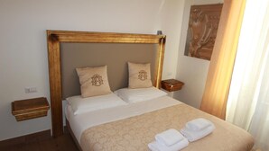 A neatly made bed with a wooden headboard, two pillows with a crest design, a bedside table with a wooden box, and a window with curtains. - Art Atelier Accomodation Camera 1 (ROMA)