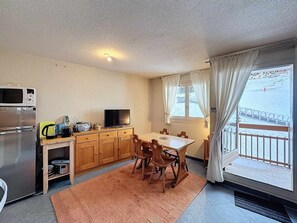 A compact kitchen with a microwave, toaster, and radio, a dining table with chairs, and a view of a snowy landscape through a balcony.