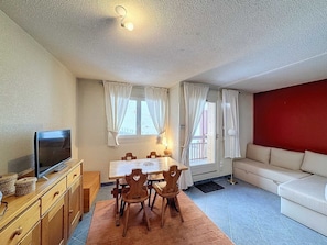 A cozy living room with a TV, wooden furniture, a red accent wall, and a balcony with curtains.
