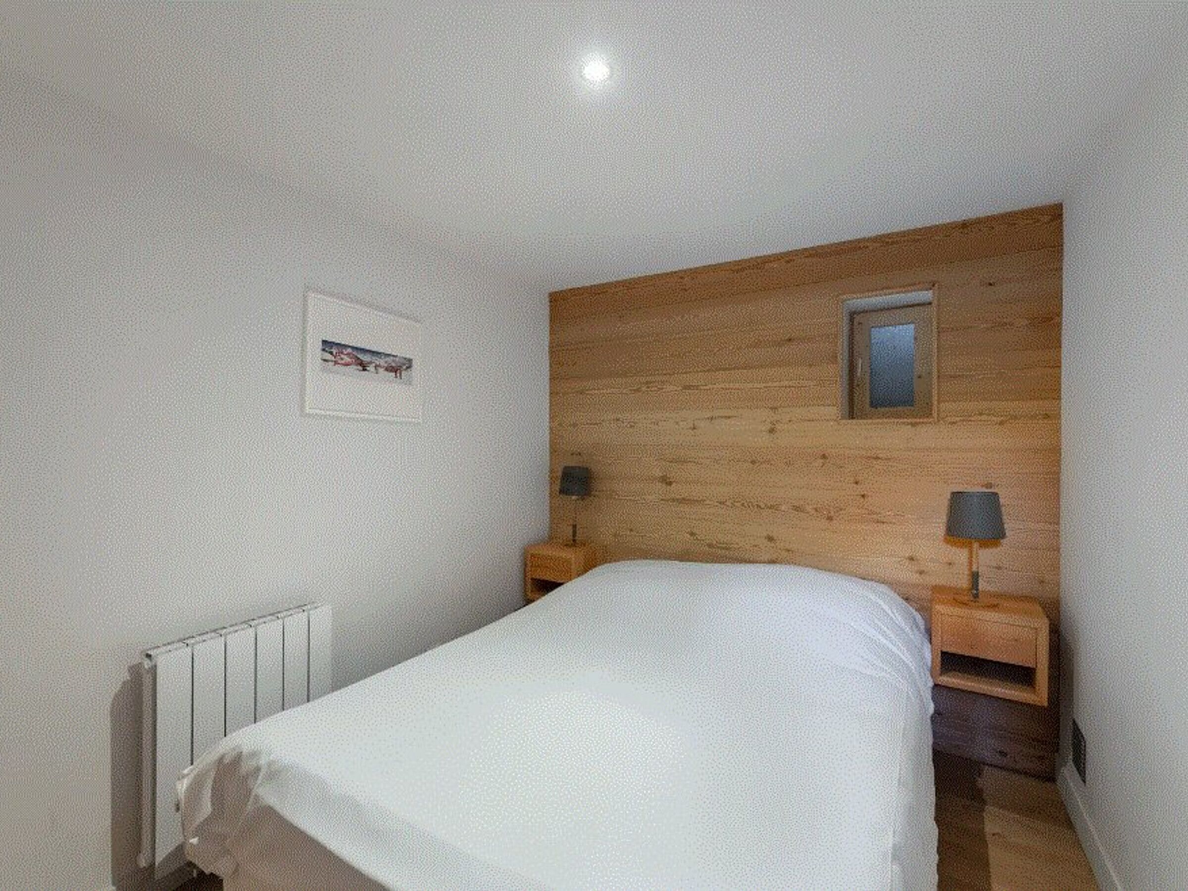 A bedroom with a wooden headboard, a single bed, two bedside tables with lamps, a small picture on the wall, and a radiator.