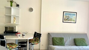 Un ambiente compatto con angolo cottura, un'area pranzo con tavolo e sedie, un divano con cuscini e una sveglia appesa al muro.