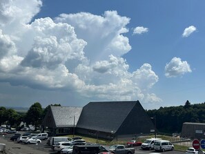A building with a sloped roof, surrounded by parked cars and a clear sky with clouds. - Apartment on the slopes in Super Besse (Besse-et-Saint-Anastaise-Super Besse)