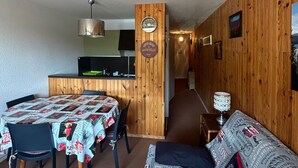 A cozy room with a wooden wall, a dining table with a quilted tablecloth, a sofa, and a kitchen area with a wooden counter and a clock. - Apartment on the slopes in Super Besse (Besse-et-Saint-Anastaise-Super Besse)