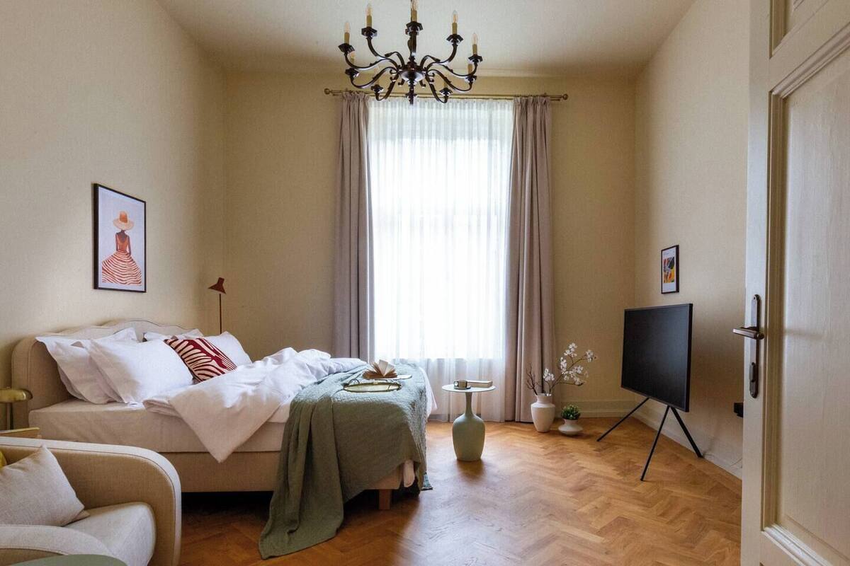 Un dormitorio con cama, televisor, candelabro, ventana con cortinas y un cuadro en la pared.