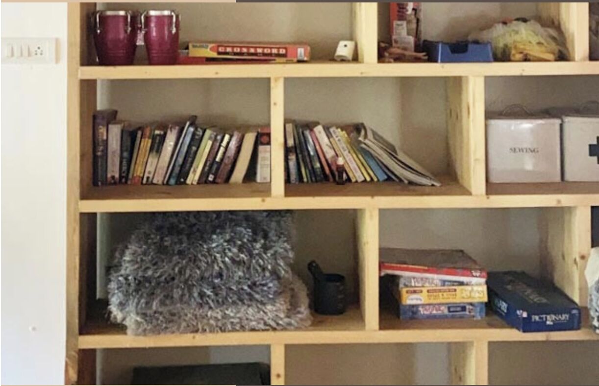 A wooden bookshelf with various items including books, a crossword book, a sewing box, and a small furry object.