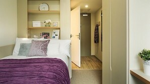 A bedroom with a bed, a bedside table, a shelf with a clock, and a door leading to another room. - Liv Student Dublin (Dublin)