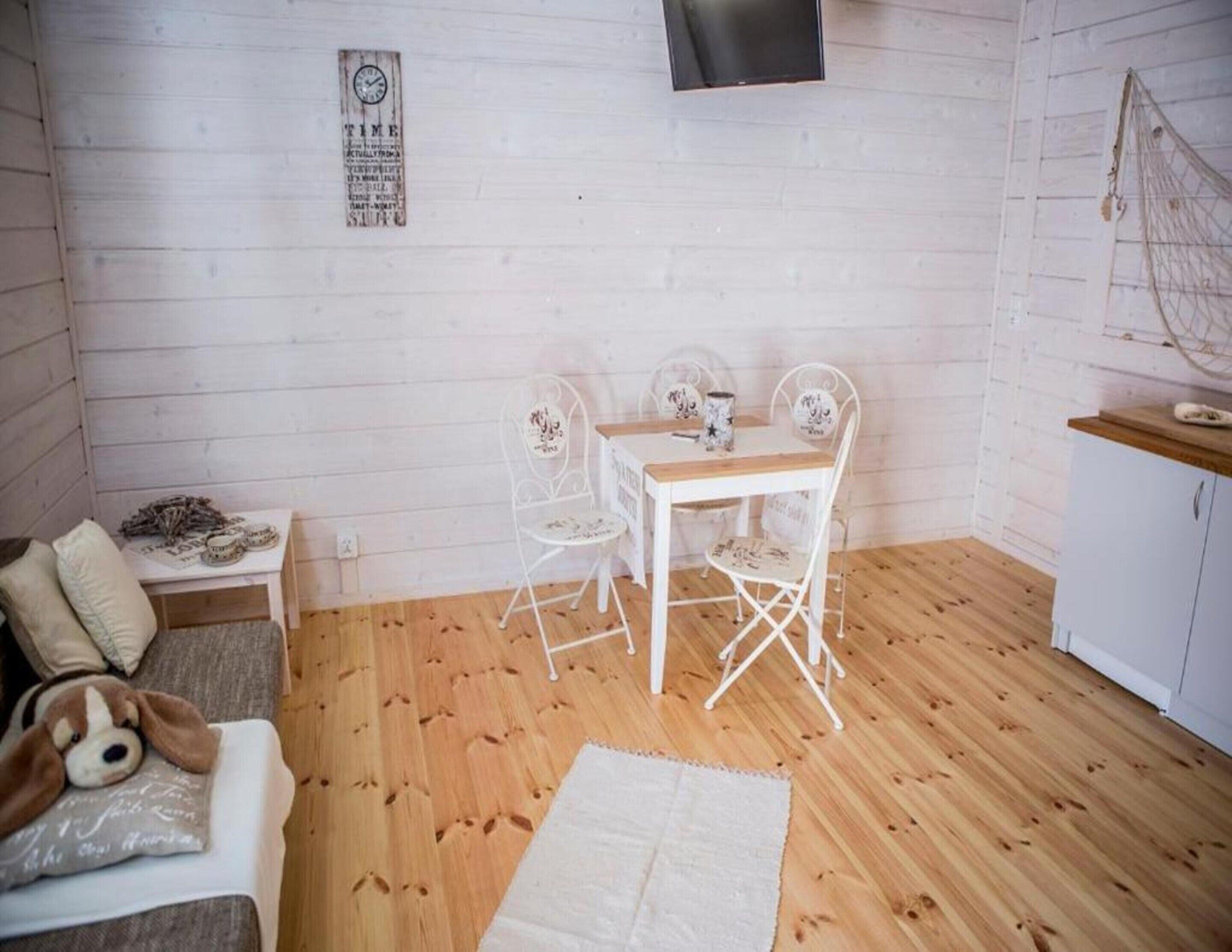 A cozy room with a wooden floor, a white sofa with cushions, a small table with a decorative item, and a white table with chairs.