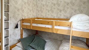 Uma cama beliche com estrutura de madeira, um beliche inferior com colchão acolchoado e travesseiros. Há uma estante à esquerda com compartimentos de armazenamento.