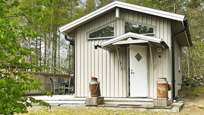 Uma cabana pequena e térrea com telhado em duas águas, uma porta de entrada e dois recipientes metálicos grandes no alpendre.