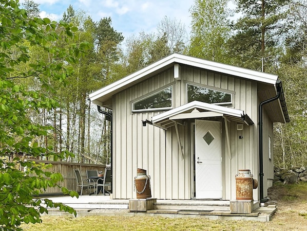 Uma cabana pequena e térrea com telhado em duas águas, uma porta de entrada e dois recipientes metálicos grandes no alpendre.