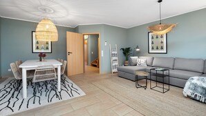 A modern living room with a dining area, featuring a white table, wicker chairs, a grey sofa, and a patterned rug.