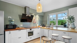 A modern kitchen with white cabinets, a wooden countertop, a built-in oven, a microwave, and a hanging light fixture. There are three bar stools and a window with a view of greenery outside.