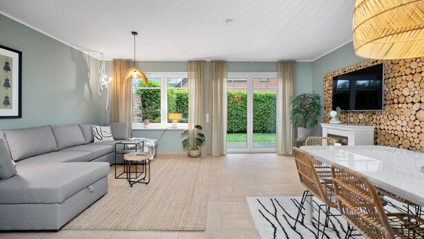 A modern living room with a grey sofa, a dining table with wicker chairs, and a TV mounted on a log-patterned wall.