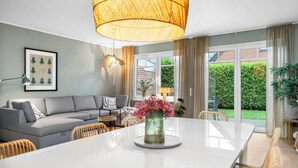A modern living room with a grey sofa, a round dining table, wicker chairs, and a large window with a view of a brick wall and greenery.