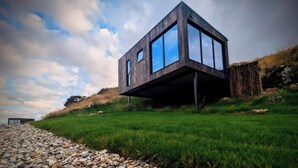 Une maison moderne, surélevée, dotée de grandes fenêtres, située sur une colline verdoyante, et accessible par un chemin de gravier.