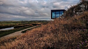 Un bâtiment moderne doté de grandes fenêtres, situé sur une colline verdoyante et offrant une vue sur une vallée et des montagnes lointaines.