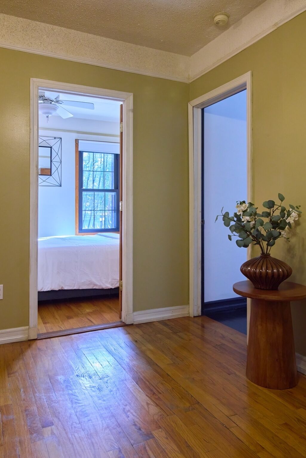 A room with wooden flooring, a bed, a window, and a vase with flowers.