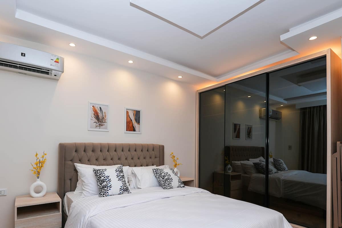 A modern bedroom with a tufted headboard, a large bed with white bedding, two wall-mounted air conditioning units, and a sliding glass door closet.