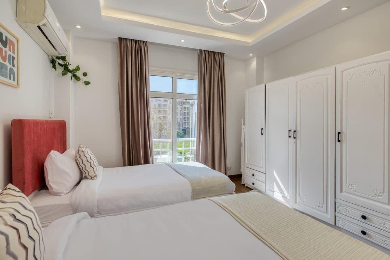 A hotel room with two beds, a large wardrobe, a window with curtains, and an air conditioning unit.