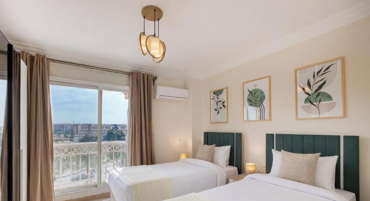 A hotel room with two beds, a balcony with curtains, a ceiling light fixture, and framed botanical artwork on the wall.