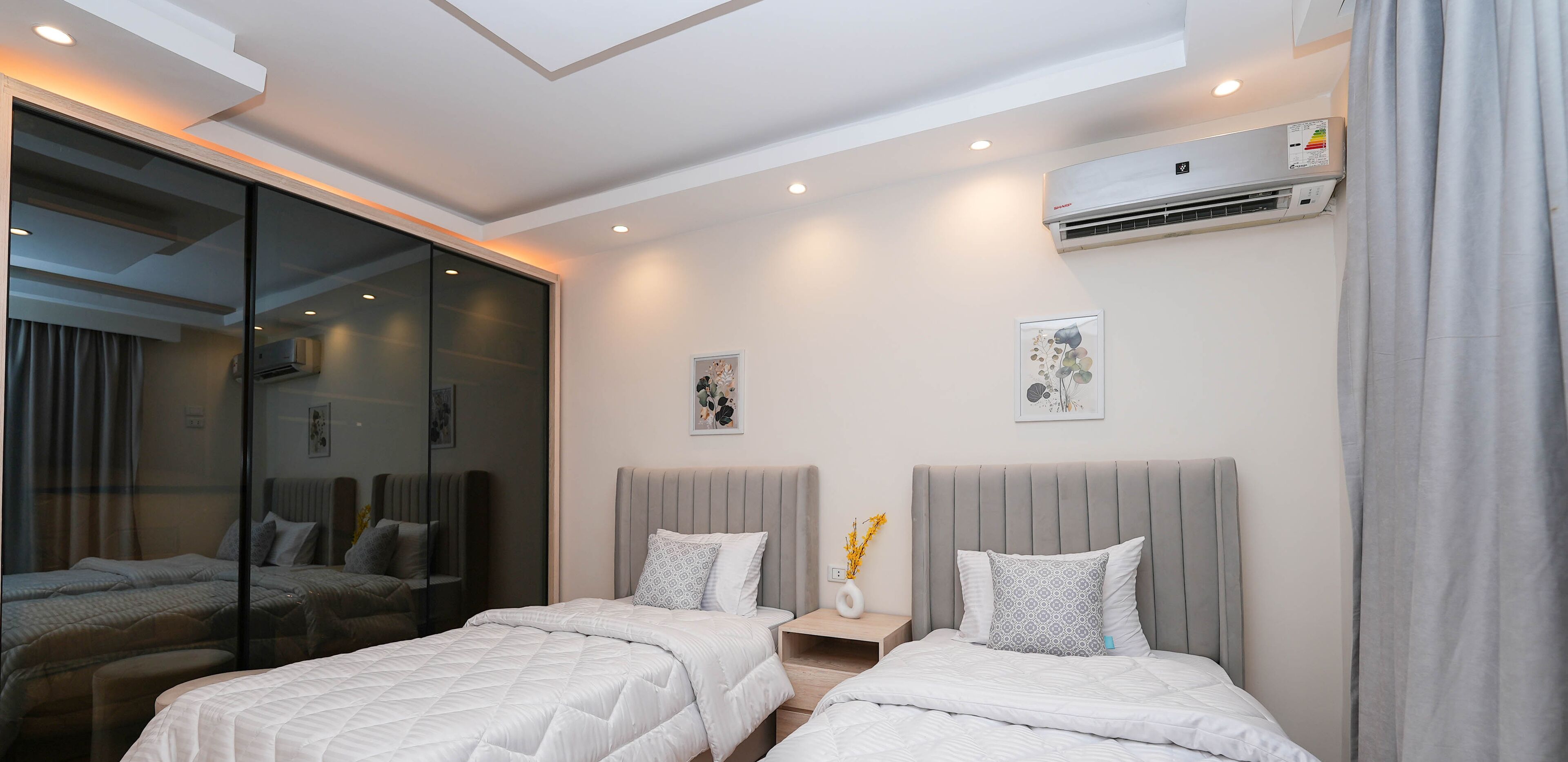 A modern hotel room with two beds, a glass partition, an air conditioning unit, and a small bedside table with a vase.