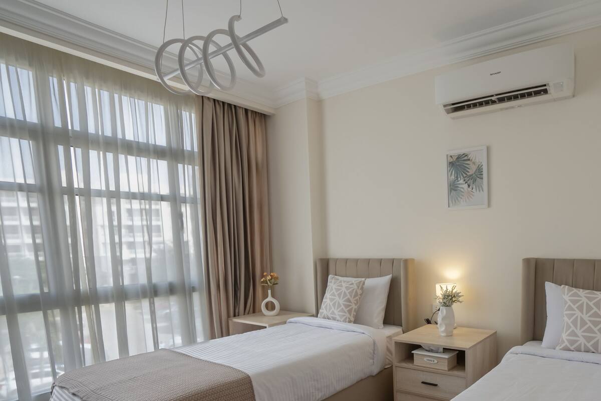 A hotel room with two beds, a large window with curtains, an air conditioner, and a wall-mounted artwork.
