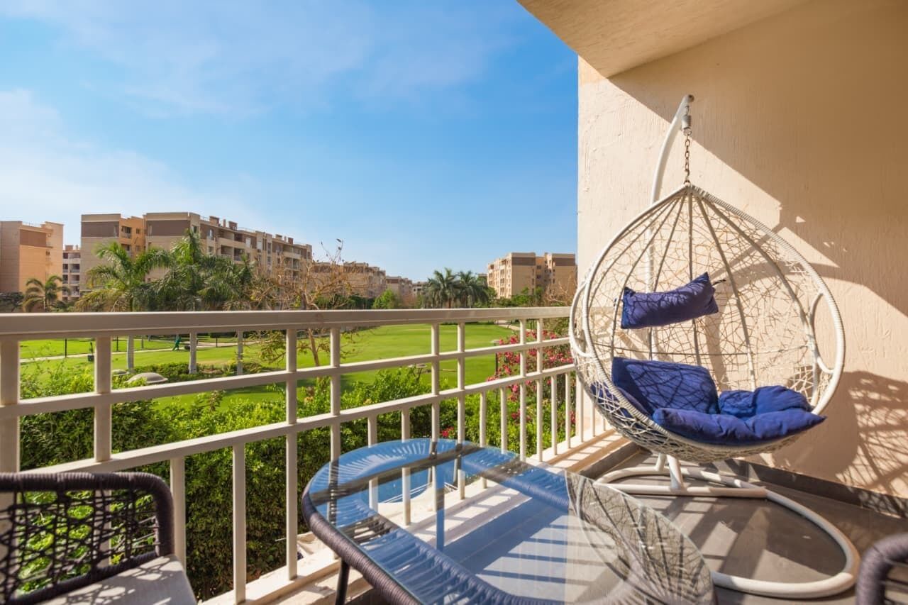 A balcony with a hanging chair, a cushioned chair, and a view of a green landscape with buildings in the background.