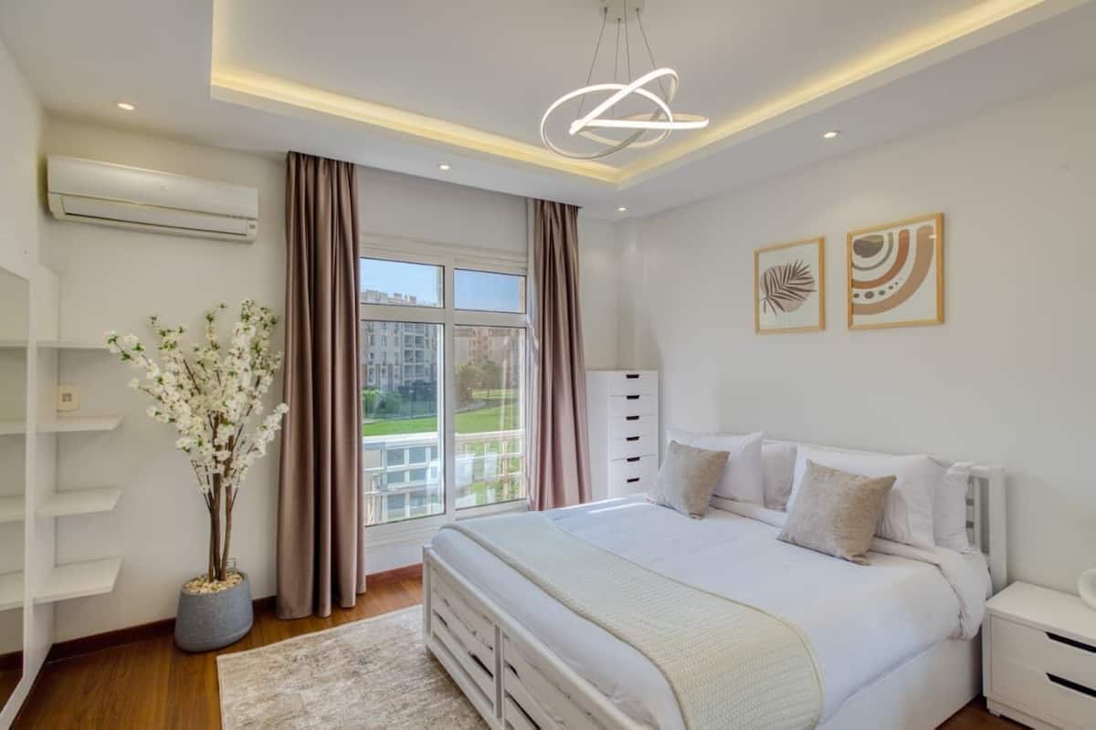 A bedroom with a large bed, a bedside table, a nightstand, an air conditioning unit, a window with curtains, and a potted plant.