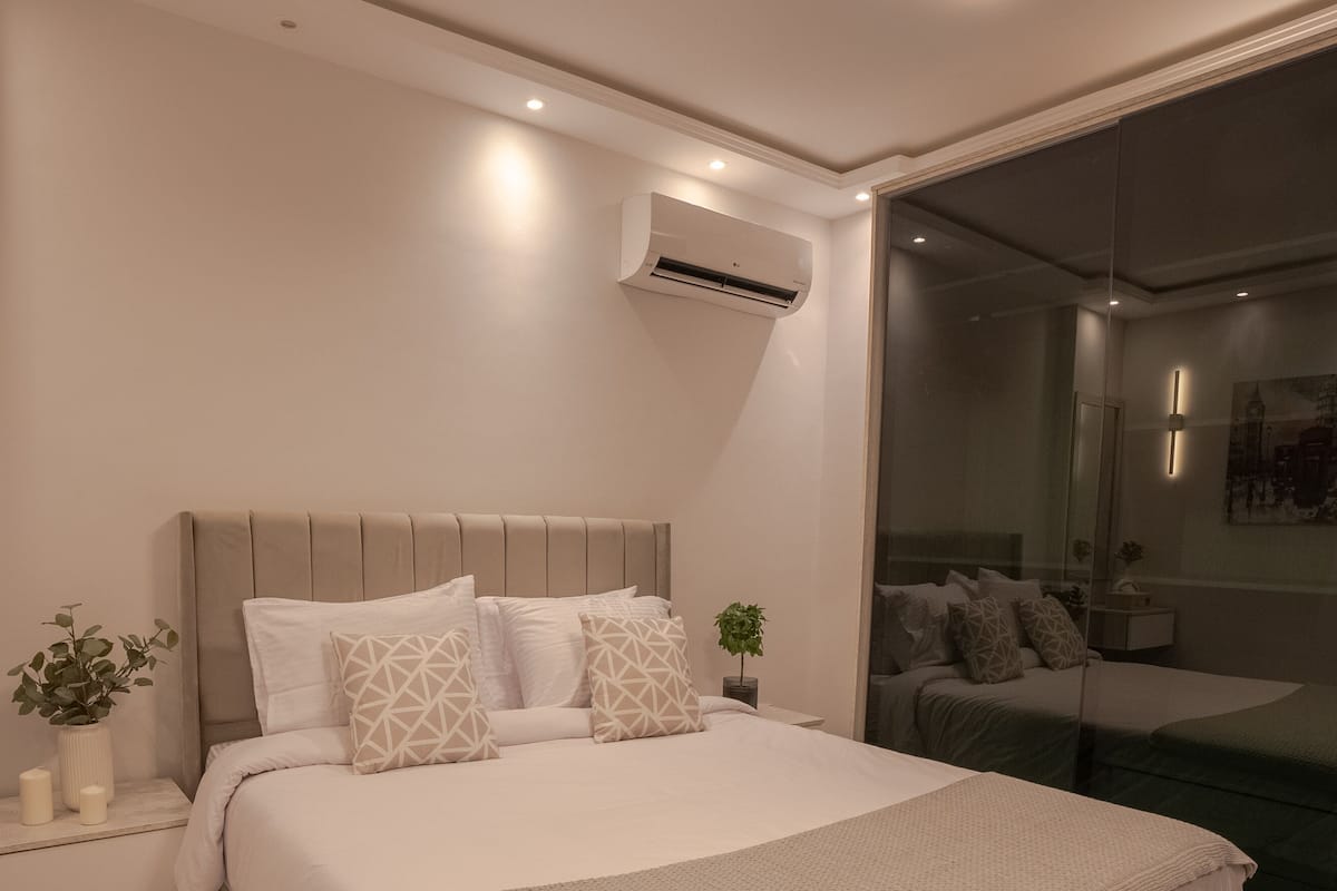 A modern bedroom with a large bed, bedside tables, a mirror, and an air conditioning unit.