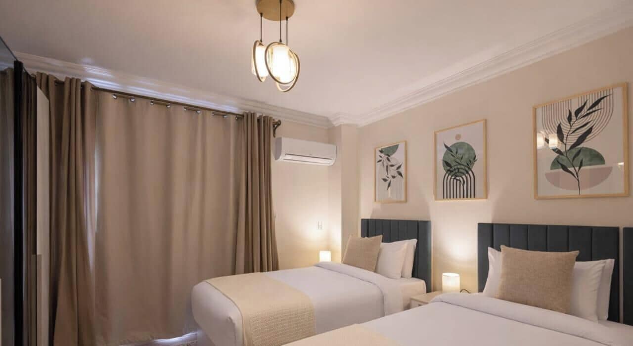 Ein Hotelzimmer mit zwei Betten, beigen Vorhängen, Wandklimaanlage und botanischen Drucken in Rahmen.