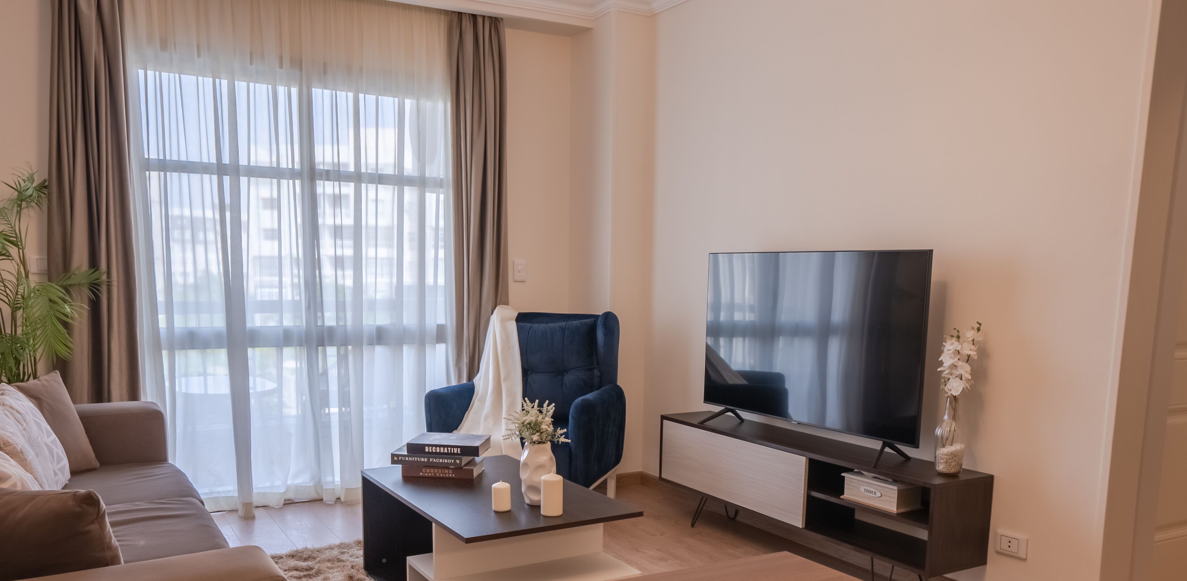 Un salon moderne comprenant un canapé, un fauteuil bleu, une table basse agrémentée d’objets décoratifs, un téléviseur posé sur un meuble et une grande fenêtre ornée de rideaux légers.