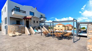 Un bâtiment moderne avec une façade en pierre, un espace extérieur abrité avec des tables et des chaises, et une vue sur la mer.