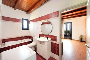 A bathroom with a bathtub, a sink, and a mirror. There is a window and a door leading to another room.