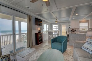 A spacious living area with a large sliding glass door to the beach, a ceiling fan, and a kitchen area.