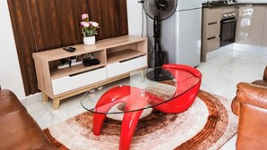 Una sala moderna con una mesa de centro de vidrio, un mueble bajo para la televisión de madera, una silla roja y una cocina al fondo.