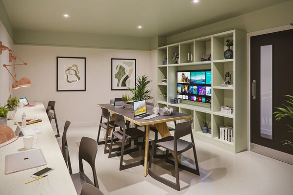 A modern office space with a long table, chairs, a laptop, a smartphone, and a desktop computer. There are two framed abstract paintings on the wall, a bookshelf with various items, and a door with a window. - Briggate Studios - Campus Accommodation (Leeds)