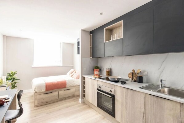 A modern kitchen with a built-in oven, a sink, and a countertop with kitchen utensils. There is a bed with a pink blanket in the adjacent room. - Briggate Studios - Campus Accommodation (Leeds)
