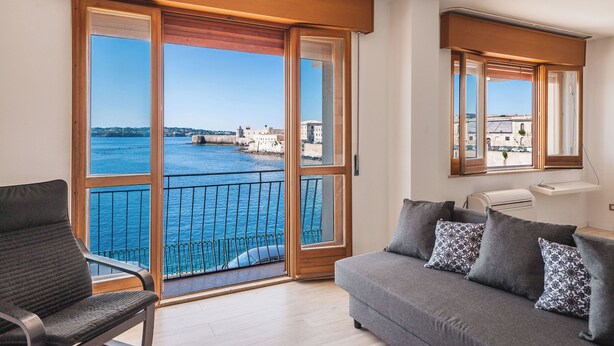 A balcony with a view of the sea and buildings, a sofa with patterned cushions, and a chair. - Una Finestra Su Cala Rossa by Wonderful Italy (Syracuse)