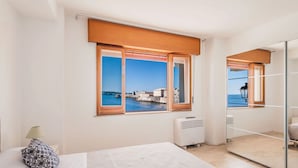 A hotel room with a large window offering a view of a coastal city, a bed with a patterned pillow, and a mirror reflecting part of the room. - Una Finestra Su Cala Rossa by Wonderful Italy (Syracuse)