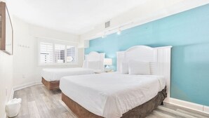 A hotel room with two beds, a window with blinds, a white nightstand, and a blue accent wall. - Oceanfront Condo Balcony with Resort Pool Hot Tub (North Myrtle Beach)