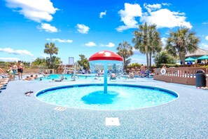Un espace piscine agrémenté d’une fontaine centrale, entouré de palmiers et surplombé d’un ciel d’un bleu limpide.