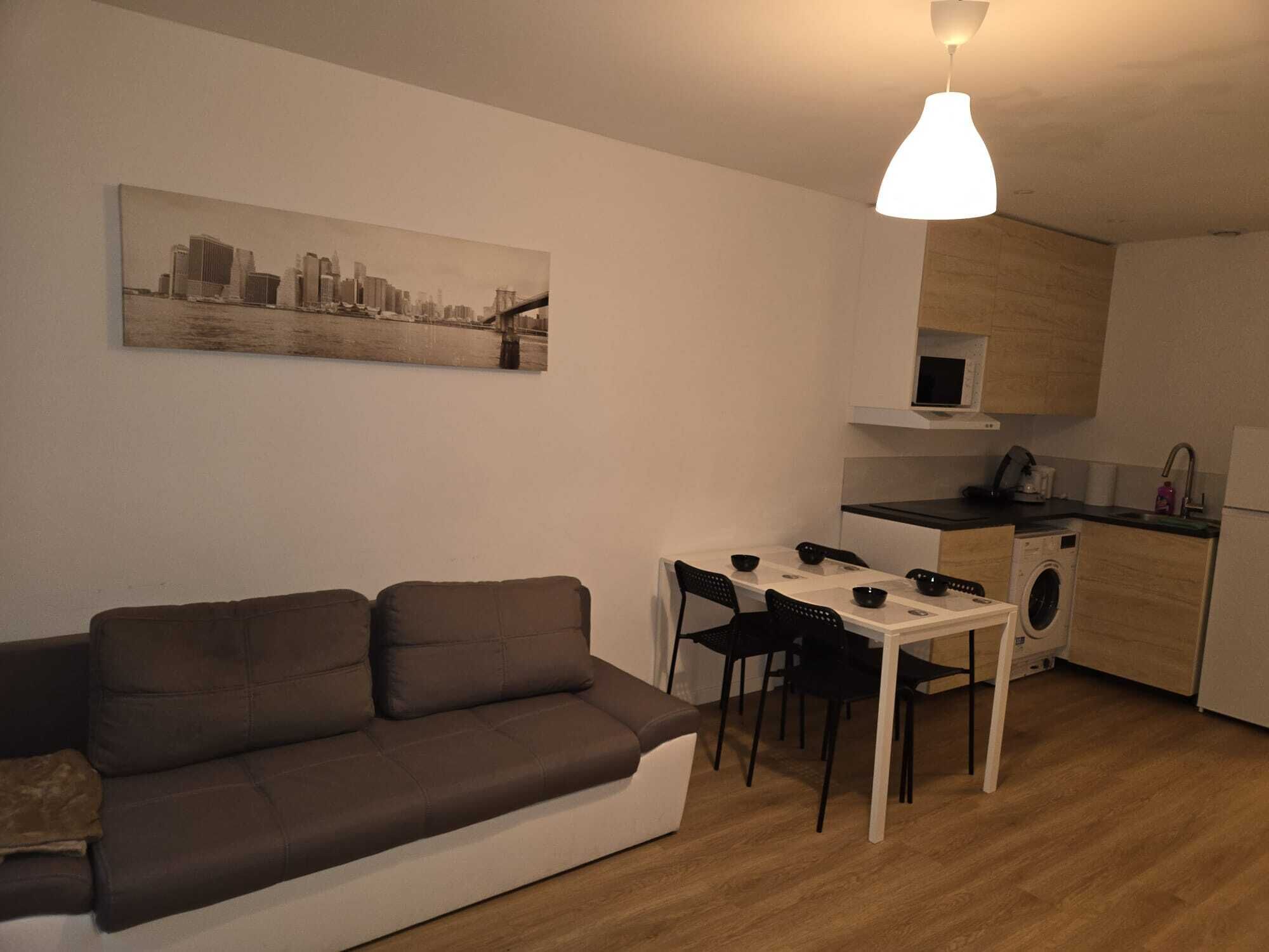 Een ruimtelijke woonkamer met een bank, eettafel, stoelen, een wasmachine en een keukenhoek.