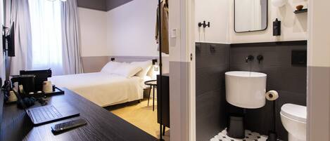 A hotel room with a bed, a desk with a laptop and phone, a bathroom with a sink and toilet, and a mirror.