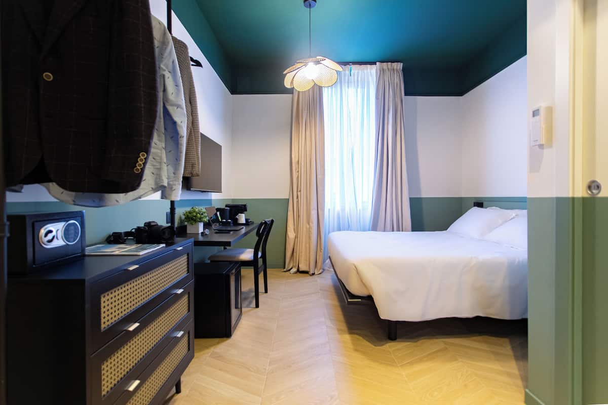 A hotel room with a bed, a desk with a chair, a dresser, and a hanging coat rack.