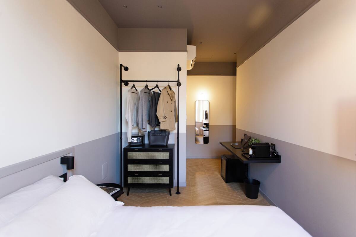 A hotel room with a bed, a wardrobe with hanging clothes, a desk with a lamp, and a small plant.