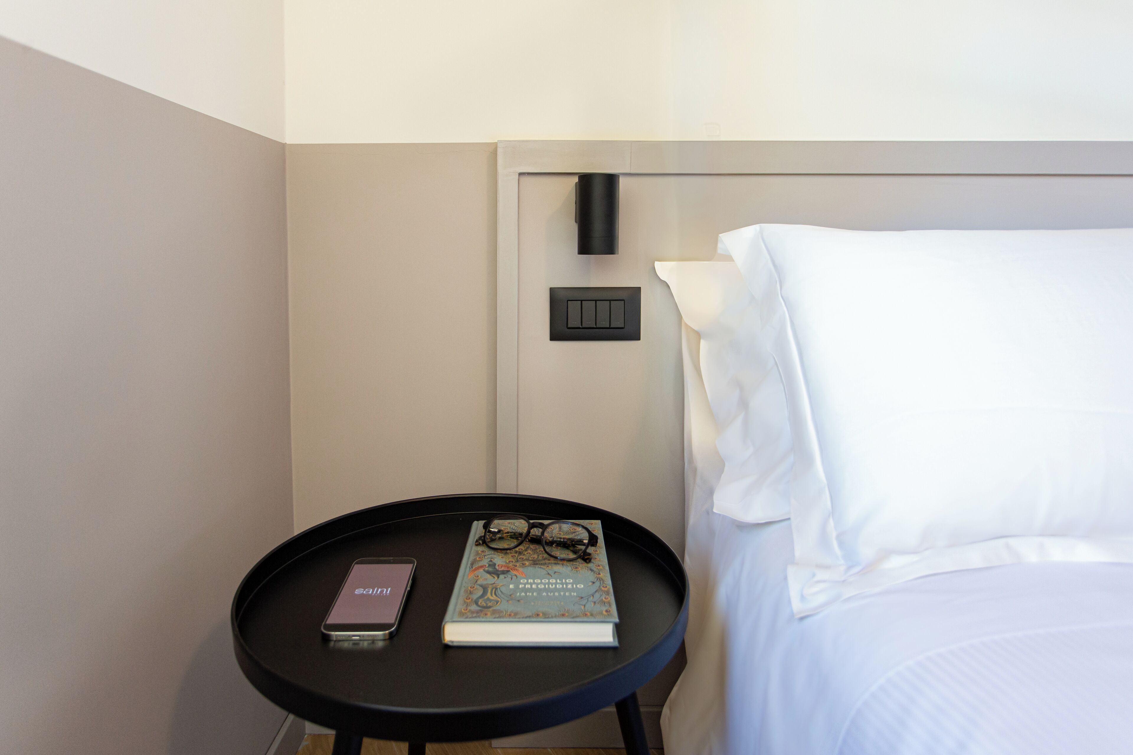 Habitación de hotel con una cama, una mesita de noche con un libro y gafas, un teléfono e interruptor de luz en la pared.